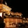 A luxurious casino at night, illuminated with warm, golden lights. The building features elegant architecture with columns and decorative elements. A few people are walking towards the entrance, and there are a couple of parked cars nearby.