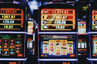A row of colorful slot machines lit with bright neon lights, showcasing various graphical elements on the screens, indicating different games and jackpots. The atmosphere suggests a lively casino setting, bustling with excitement.