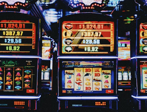 A row of colorful slot machines lit with bright neon lights, showcasing various graphical elements on the screens, indicating different games and jackpots. The atmosphere suggests a lively casino setting, bustling with excitement.