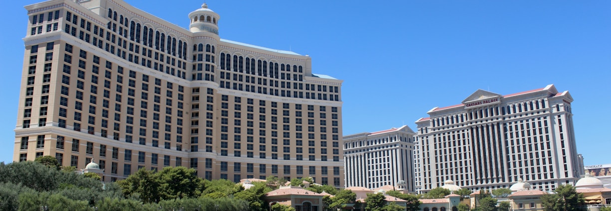 A large luxury hotel and casino building with multiple stories, featuring a light facade with arched windows. The structure has a prominent central tower with a dome-like top. It is surrounded by landscaping with trees and a body of water in the foreground.