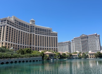 A large luxury hotel and casino building with multiple stories, featuring a light facade with arched windows. The structure has a prominent central tower with a dome-like top. It is surrounded by landscaping with trees and a body of water in the foreground.